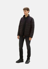 Czarna kurtka puchowa z zapięciem na zamek i wysokim kołnierzem, z pikowanymi sekcjami i kieszenią na zamek, noszona na czarną koszulę i dżinsy.