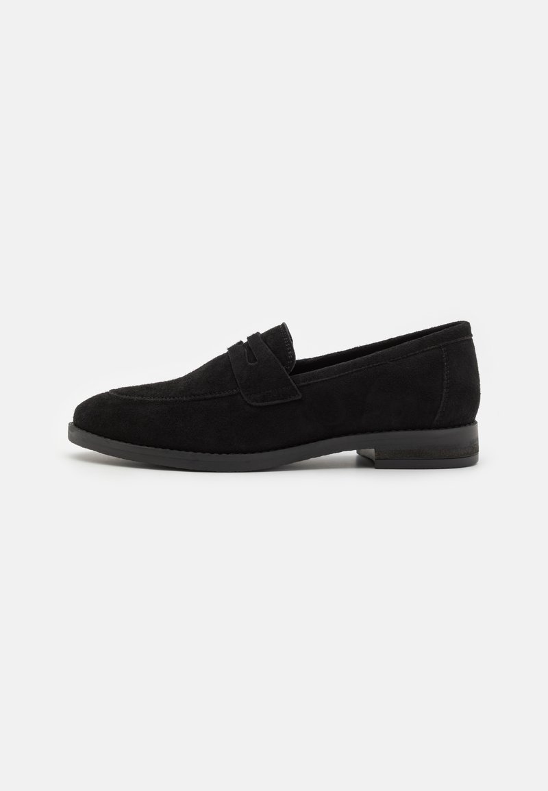 Mocassin en daim noir avec un petit talon, un bout arrondi et un détail de bride sur le cou-de-pied, vu de profil sur un fond blanc.