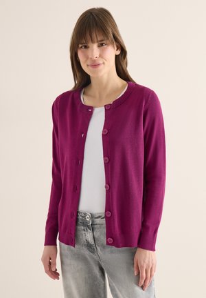 Femme portant un cardigan rose foncé boutonné, une chemise blanche et un jean gris, debout devant un fond beige uni.