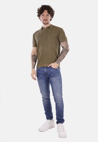 Polo shirt a righe verde oliva con maniche corte, abbinato a jeans blu slim-fit e sneakers bianche. Presenta tatuaggi sulle braccia.