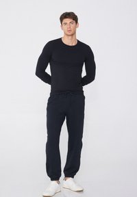 Schwarzes Langarmshirt kombiniert mit dunkelblauen Jogginghosen, die einen elastischen Bund, seitliche Taschen und gerippte Bündchen haben. Kombiniert mit weißen Sneakers.