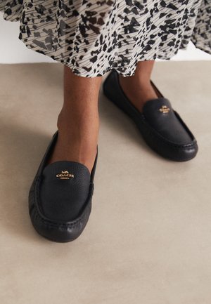 Fødder iført sorte læder Coach loafers kombineret med et hvidt og sort mønstret skørt på en beige gulv.