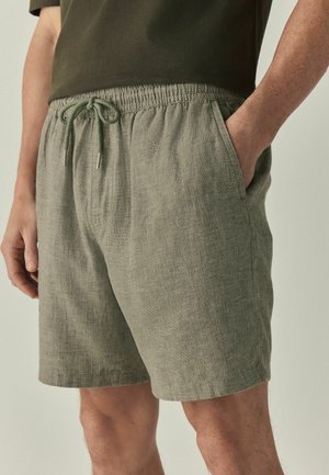 Mann trägt olivgrüne Leinen-Shorts mit Kordelzug und Taschen, kombiniert mit einem dunkelolivfarbenen Kurzarmhemd, steht mit einer Hand in der Tasche.