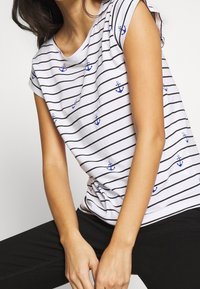 Vit T-shirt med svarta horisontella ränder och blåa ankarmönster. Kort ärm, lös passform. Bärs med svarta byxor. Material i bomull.