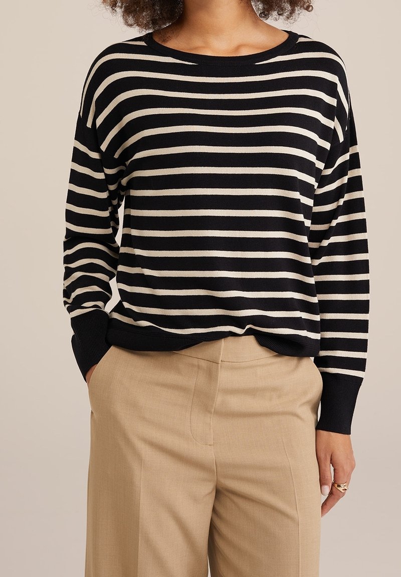 Femme portant un haut à manches longues rayé noir et blanc, rentré dans un pantalon beige taille haute, les mains dans les poches.