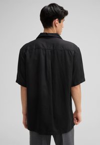 Schwarz kurzärmeliges Shirt aus leichtem Stoff. Mit klassischem Kragen, lässigem Schnitt, geradem Saum und einem Rückenfalten.
