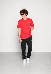 Rød bomulds-T-shirt med Nike-logo, parret med sorte sweatpants med et logo, og hvide sneakers. Model iført solbriller.