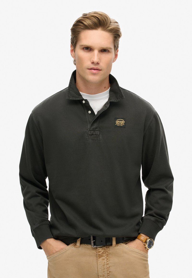 Dark green long-sleeve polo shirt with a collared neck and embroidered logo on the chest. Worn with neutral-colored pants and a black belt.