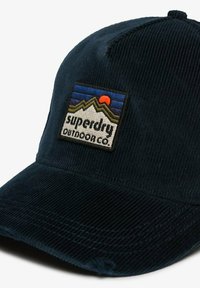 Gorra de pana azul oscuro con una forma estructurada, que presenta un parche bordado con montañas y un sol, etiquetado como "superdry outdoor co."