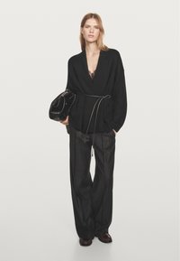 Černý oversized kardigan s překládaným designem, zavazovaný v pase. Kombinován s volnými pruhovanými kalhotami a lesklou černou kabelkou.