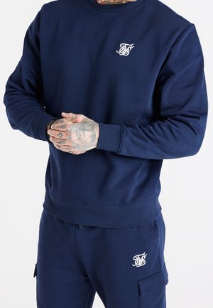 Mann trägt dunkelblauen Sweatshirt und passende Cargohose mit weiß gesticktem Logo auf Brust und Oberschenkel, Hände verschränkt, zeigt Tattoos.
