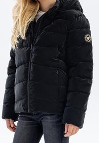 Sort dunjakke med hætte, med quiltede sektioner, en frontlynlås og et rundt logo-patch på venstre ærme.