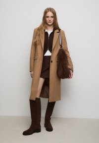 Cappotto in misto cammello con rever sartoriali, stivali alti fino al ginocchio in suede marrone, giacca marrone con zip e borsa in pelliccia sintetica marrone con tracolla.