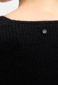 Gros plan sur le haut du dos d'une personne portant un pull noir texturé avec une petite étiquette ronde en argent attachée près de l'encolure.