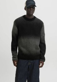 Mann trägt einen schwarz-grau verlaufenden Strickpullover, dunkle weite Jeans und eine schwarz-beige "Pasadena" Baseballmütze.