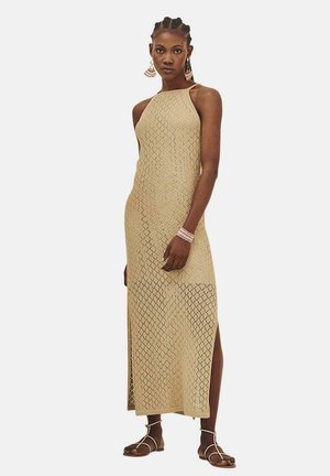 Vestido maxi de punto en color beige claro con un patrón de diamantes, aberturas laterales y un escote alto. Combinado con sandalias planas y joyería.