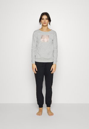 Woman standing barefoot wearing a light gray sweatshirt with a rose gold design and black jogger pants with side stripes.