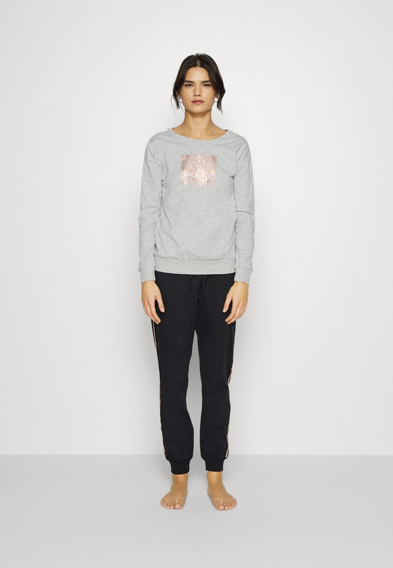 Kvinde står barfodet iført en lysegrå sweatshirt med et rosaguldsdesign og sorte joggerbukser med sidelister.