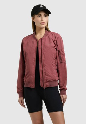 Woman wearing a dusty rose bomber jacket, black shorts, and a black baseball cap against a plain gray background.