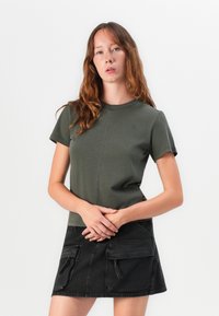 Maglietta corta olive con collo girocollo e dettagli di cucitura, abbinata a una gonna di jeans nera con tasche cargo frontali e zip.