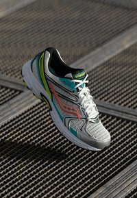 Scarpa da corsa bianca Saucony con accenti turchesi, neri, giallo neon e arancioni appoggiata su una superficie in griglia metallica testurizzata.