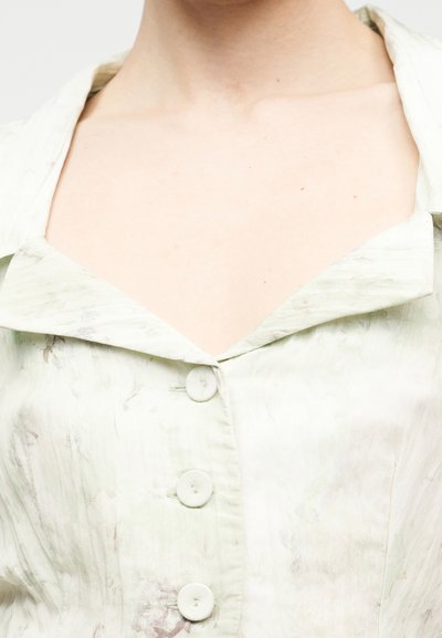 Πράσινο ανοιχτό πουκάμισο με φαρδιά γιακά, με υφή υφάσματος και αχνά, αφηρημένα μοτίβα. Τέσσερα κουμπιά εμφανή στο μπροστινό μέρος.