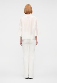 Mujer con corte bob recto, vestida con una blusa blanca transparente de mangas acampanadas y pantalones blancos de pierna ancha, de pie contra un fondo liso.
