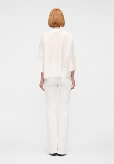 Γυναίκα με ίσιο καρέ κούρεμα που φοράει διάφανο λευκό πουκάμισο με μανίκια που ξεφύτρωναν και λευκό παντελόνι φαρδύ, στέκεται μπροστά σε απλό φόντο.