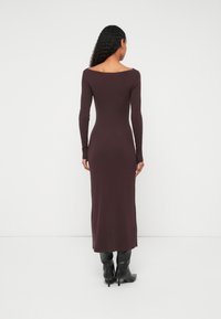 Rievota, garām piedurknēm kleita dziļā burgundijas krāsā ar apaļāku kakla izgriezumu, piegulošu siluetu un potītes garuma apakšu. Valkāta ar melniem ceļgalu augstiem zābakiem.