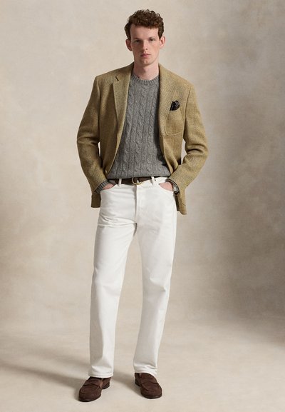 Mann trägt beigen Blazer, grauen Zopfstrickpullover, weiße Hose, braune Schuhe, steht mit Händen in den Taschen vor neutralem Hintergrund.