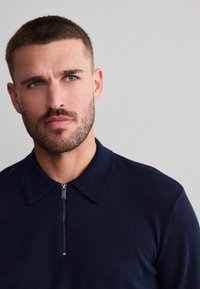 Plyšový polo svetr na zip v námořnické modři, vyrobený z měkké tkaniny, s límečkem, krátkým zipem vpředu a hladkou texturou. Minimalistický design a přiléhavý střih.