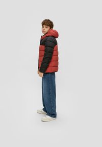 Rote und schwarze Steppjacke mit Kapuze, mit einem gesteppten Design und Reißverschluss. Kombiniert mit lockeren blauen Jeans und weißen Sneakers.