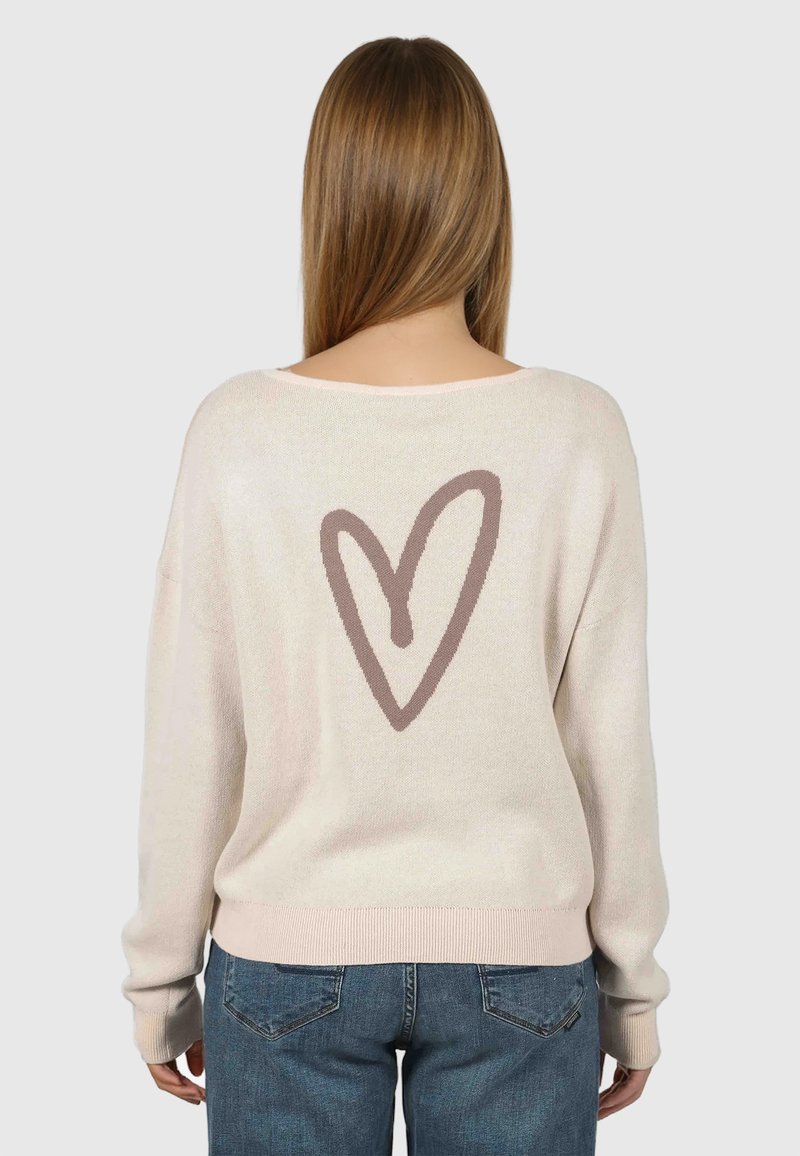 Femme aux cheveux longs portant un pull beige clair avec un grand motif en forme de cœur marron dans le dos, associé à un jean bleu.