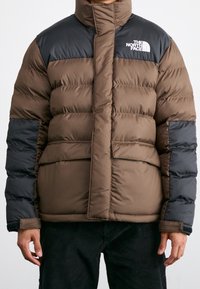 Chaqueta acolchada aislante de color marrón y negro con cuello alto, cierre de cremallera y logo en el pecho; diseño acolchado y dos bolsillos delanteros.