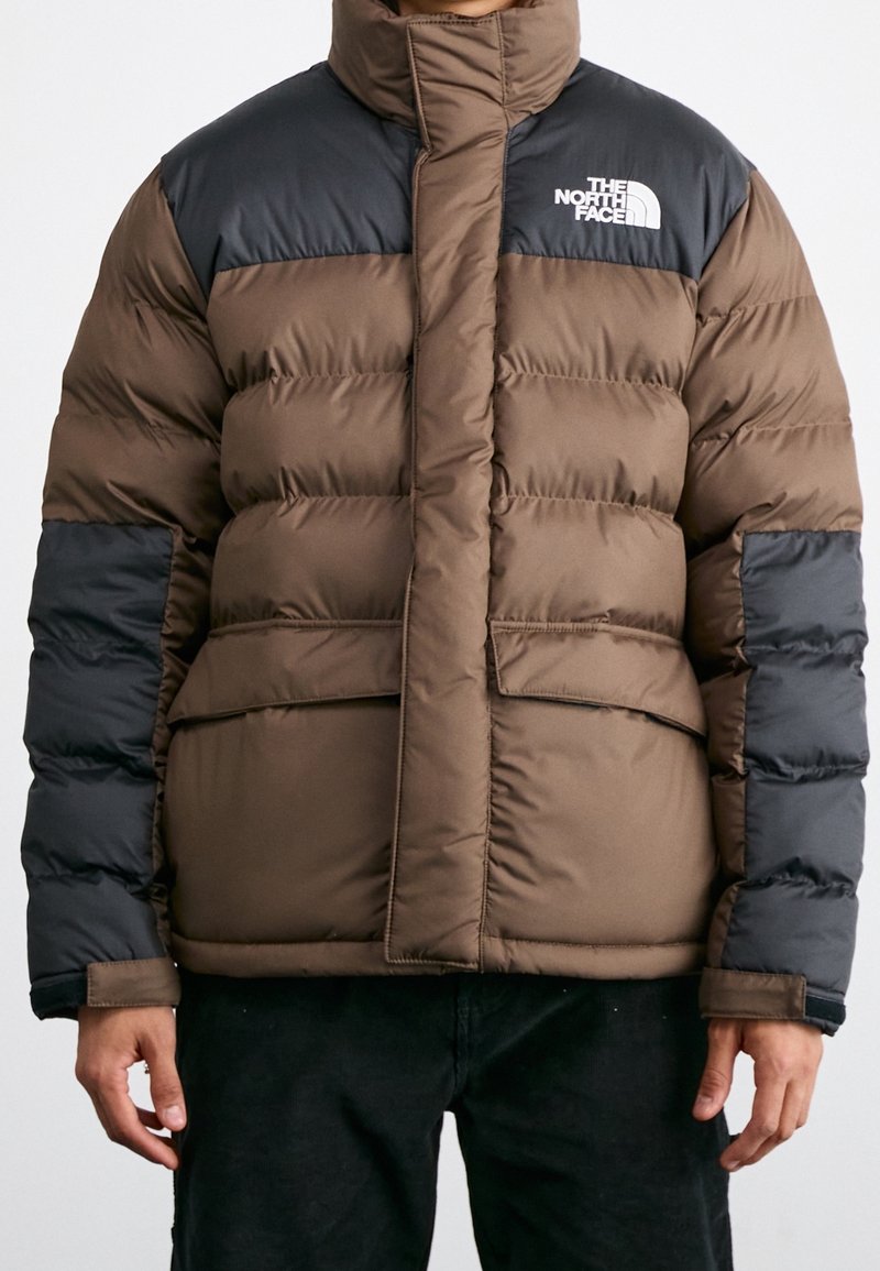 Chaqueta acolchada aislante de color marrón y negro con cuello alto, cierre de cremallera y logo en el pecho; diseño acolchado y dos bolsillos delanteros.