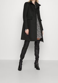 Cappotto di lana nera con colletto alto, chiusura a bottoni e tasche laterali. Indossato sopra un vestito con stampa e abbinato a stivali di pelle alti fino al ginocchio.