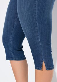 Tummansinisiä farkku capri-housuja, joissa on tiukka malli. Ominaisuuksina polvimittainen leikkaus ja pienet halkiot helmassa. Silkkinen pinta.