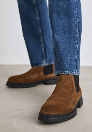 Bottines en daim marron avec panneaux latéraux élastiques noirs, portées avec un jean en denim bleu sur un sol clair.