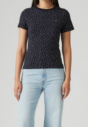 Vrouw draagt een zwart T-shirt met korte mouwen en witte stippen en een lichtblauwe jeans met hoge taille, staand tegen een effen achtergrond.