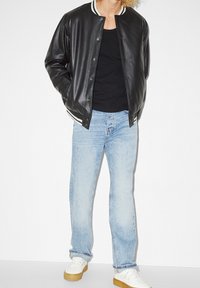 Veste bombardier en cuir noir avec col côtelé rayé blanc, associée à un jean bleu clair et des baskets blanches avec semelles en caoutchouc beige.