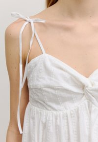 Witte jurk met dunne, verstelbare bandjes. Kenmerkt zich door een gestructureerde stof met verticale strepen en een geraffineerde corsage. Zachte en lichte materialen.