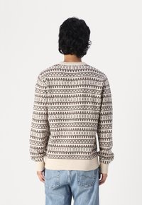 Stickad tröja i krämfärg med bruna geometriska mönster, rund halsringning, ribbade ärmslut och nederkant, med en texturerad yta.