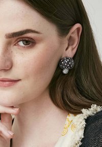 Svarta och silverfärgade runda örhängen med en texturerad yta, som har en klunga av glittrande ädelstenar vid basen, bärs på en modellens öra.