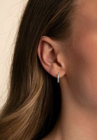 Silver hoops med en strukturerad yta och små, inbäddade klara stenar. Visas på en ljus bakgrund nära en personens öra.