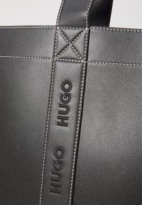 Svart läder tote-väska med synlig sömnad och präglad "HUGO"-logotyp på remmen. Har en strukturerad design med kontrasterande sömmar.