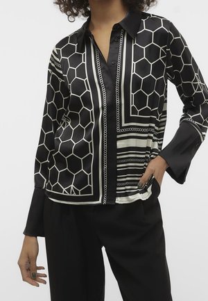 Femme portant un chemisier à manches longues noir et blanc à motifs hexagones et rayures, associé à un pantalon noir ample.
