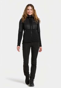 Leende kvinna som går framåt klädd i svart vadderad jacka, svarta byxor och svarta vandringskängor mot en enfärgad bakgrund.