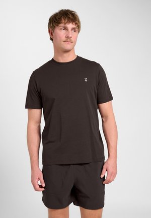 Mężczyzna w czarnym T-shircie z krótkim rękawem i czarnych szortach, stojący i lekko patrzący w bok na jednolitym jasnym tle.