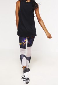 Czarna bezrękawnik zestawiona z legginsami w geometryczne wzory w odcieniach fioletu, żółci i bieli, noszona z sportowymi sneakersami.