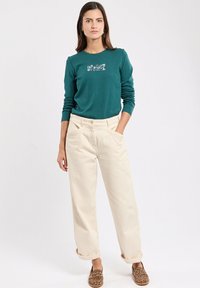 Türkises Langarmshirt mit floralem Grafikdesign, kombiniert mit beigen, lockeren Hosen und braunen Slippern im Leopardenmuster.